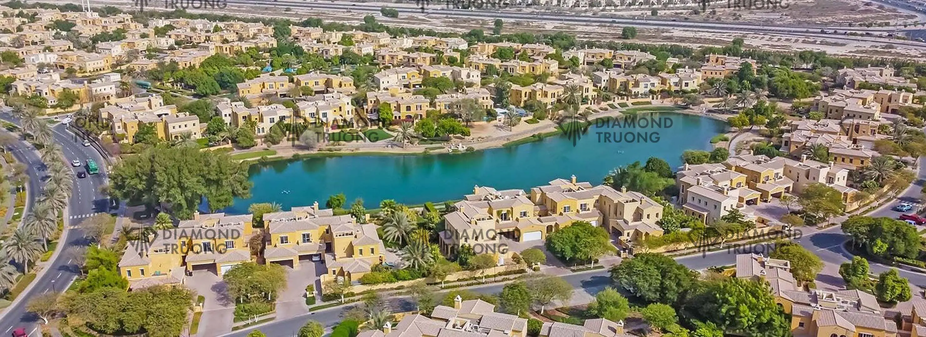 Aerial view of ochre villas with tiled roofs around a turquoise lake, with a main road on the left