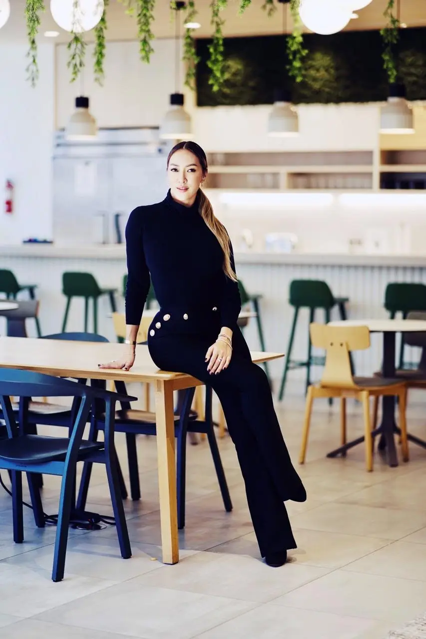 Diamond Truong in black, perched on the edge of a wooden table in a bright café hung with plants