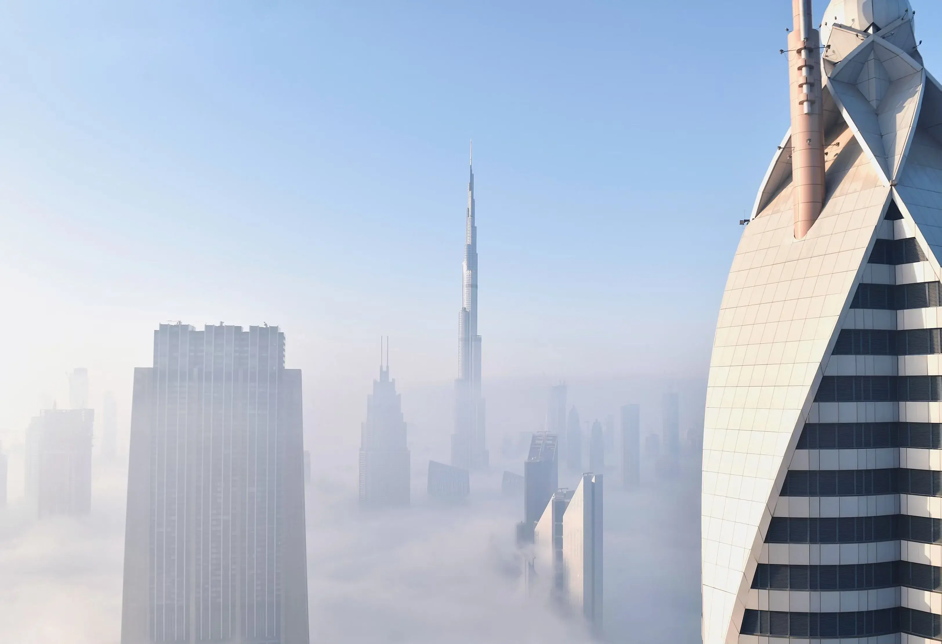 Skyscrapers emerging through thick fog with Burj Khalifa prominently in the center under clear blue sky.