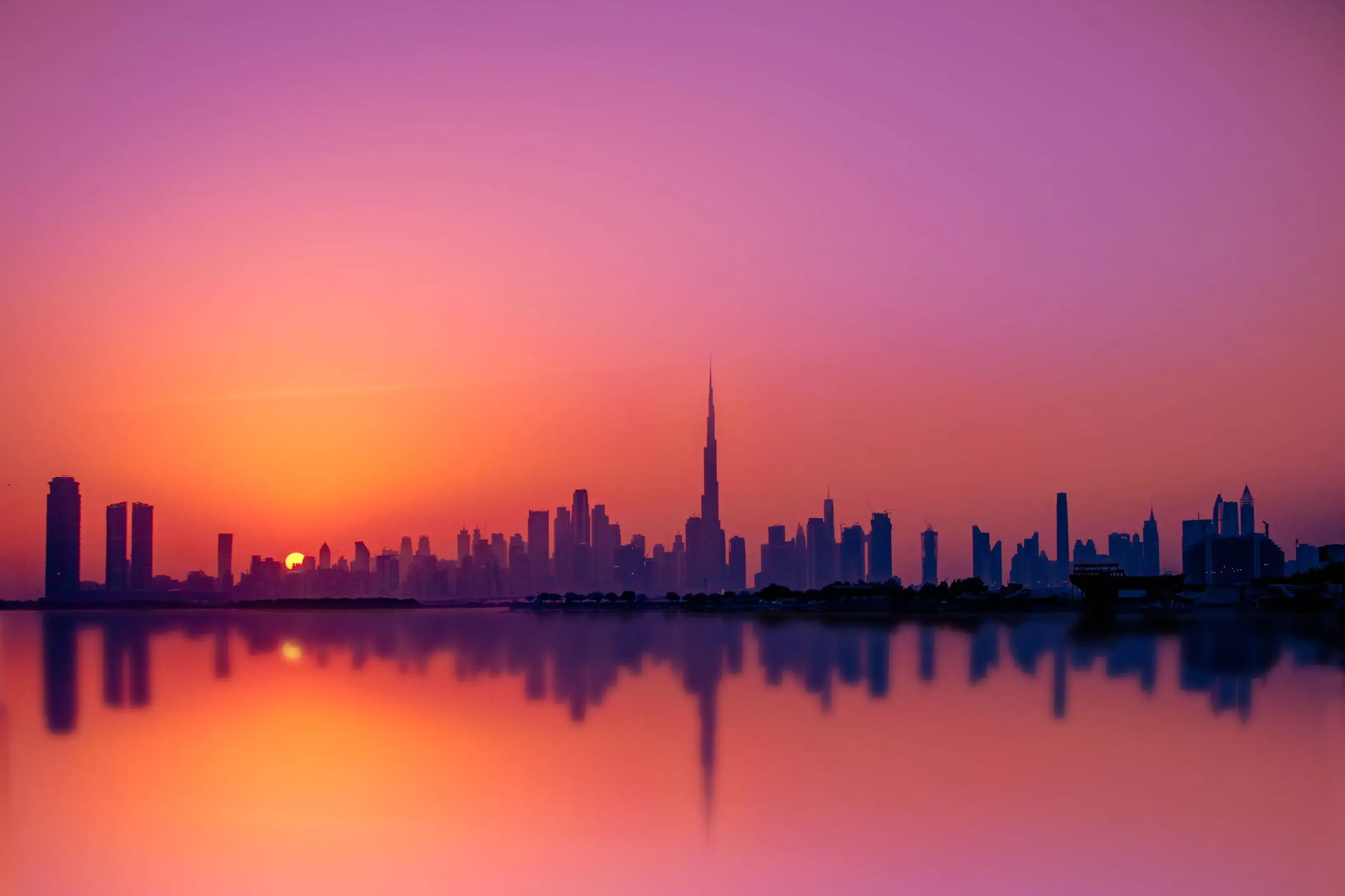 Silhouette of Dubai skyline including Burj Khalifa at sunset with colorful sky and reflection in water.