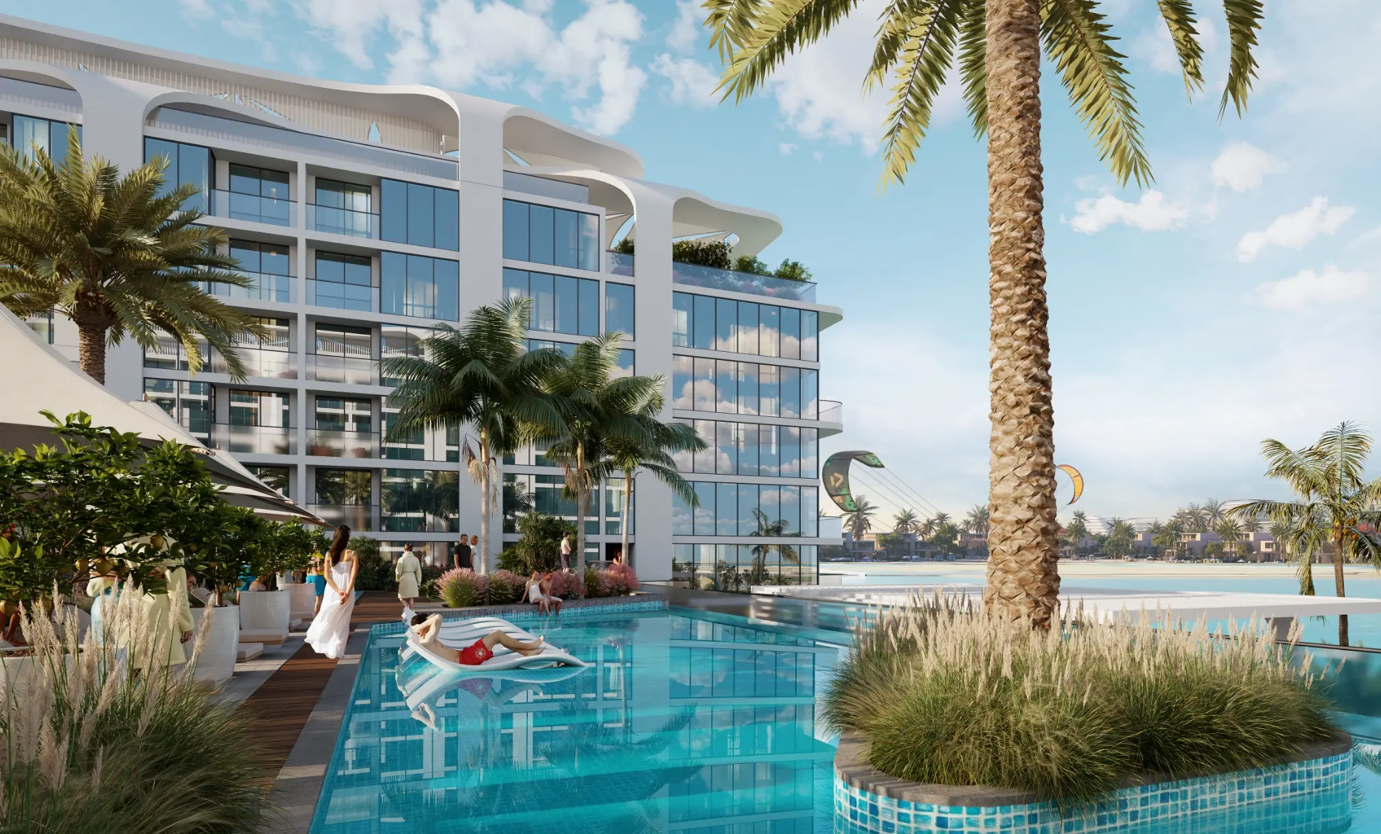 Render of a pool with an in-water lounger and a planted island under a palm, beside a white apartment block, with kite surfers beyond