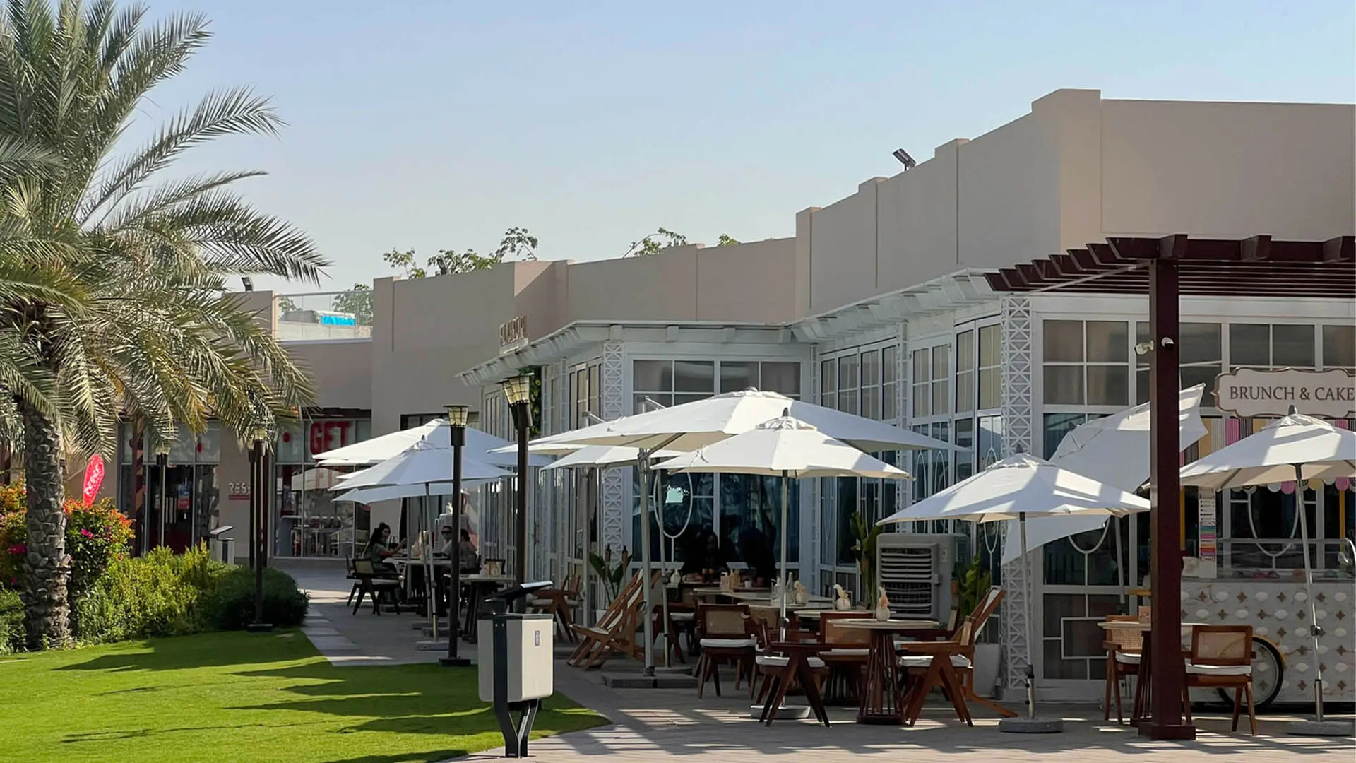Café tables under white parasols outside a low-rise row of shops and restaurants, beside palms and a lawn