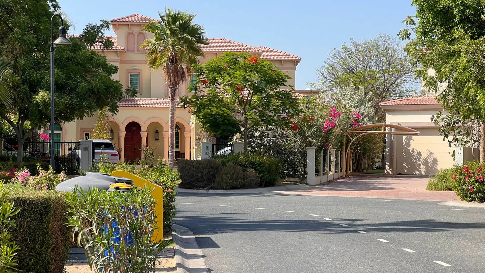 A two-storey villa with a terracotta roof and an arched porch behind flowering trees, on a quiet residential street