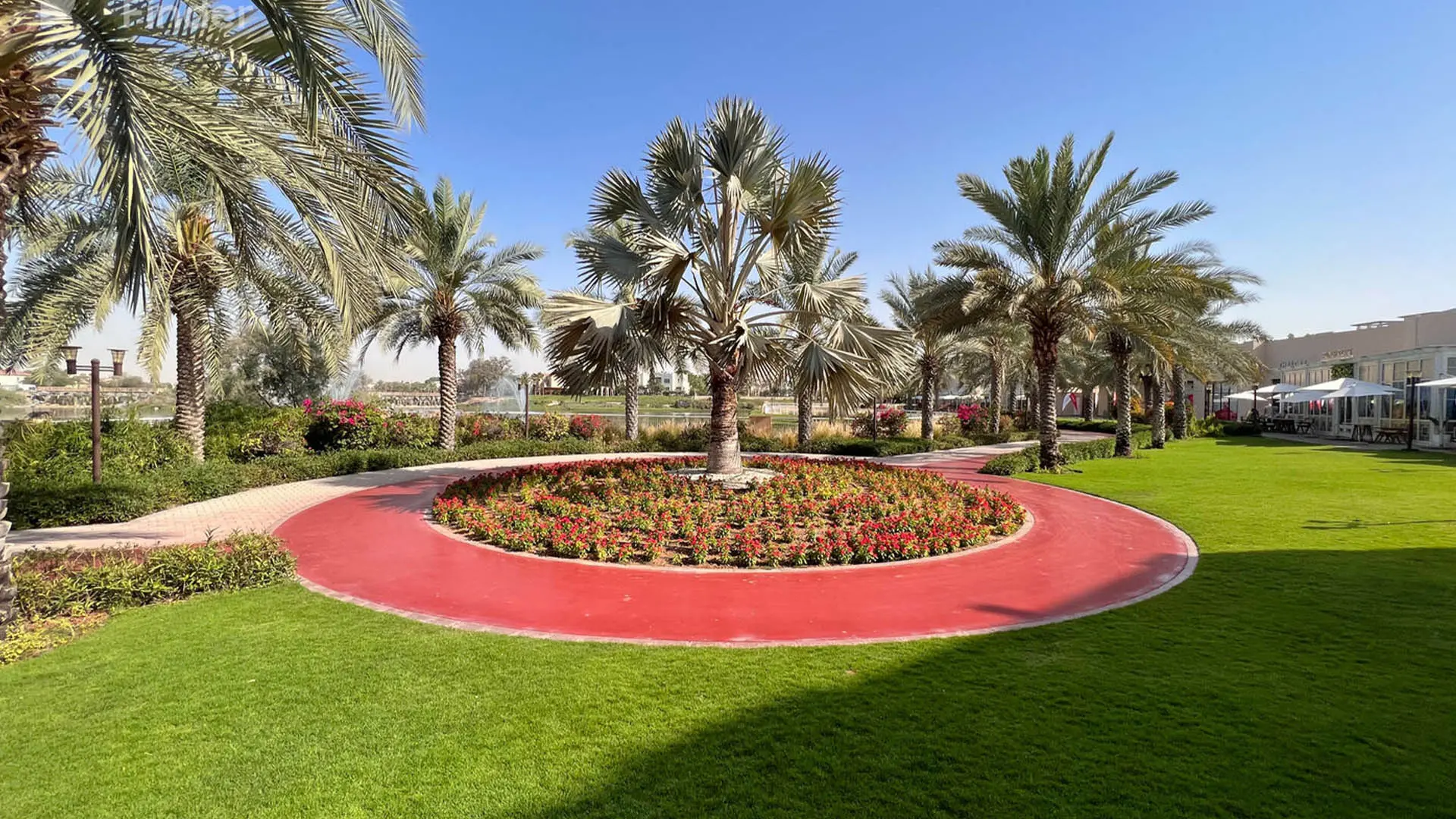 A red running track loops round a flower bed under palms, beside a lake with a fountain and a row of cafés