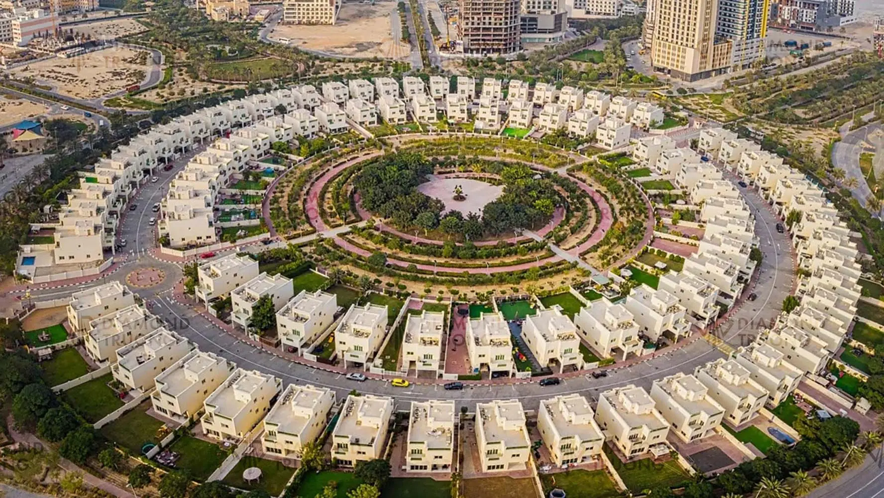 Aerial view of a ring of white three-storey villas around a planted park in Jumeirah Village Circle, with apartment towers and a crane behind