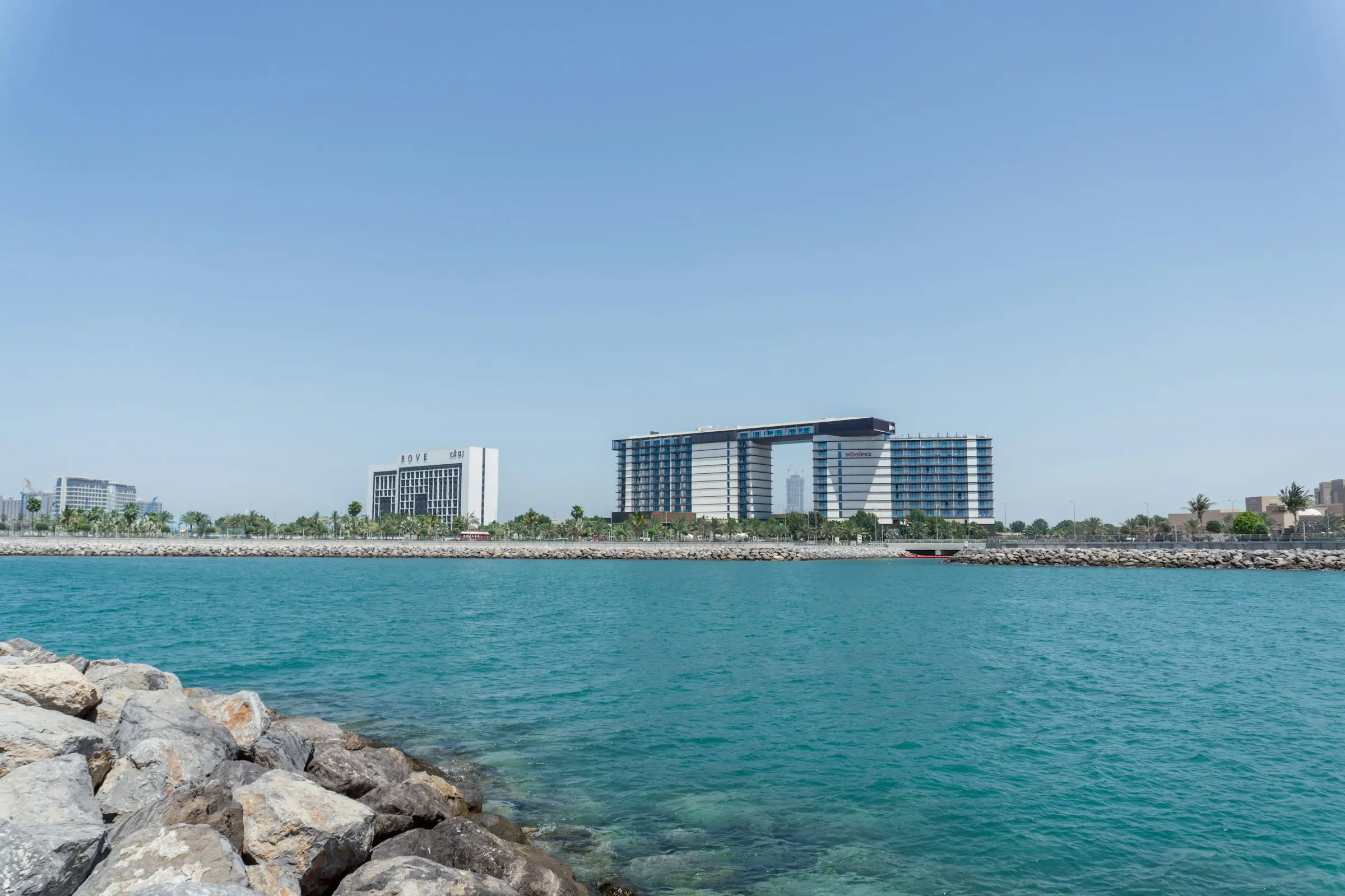 Beach hotels on Al Marjan Island across clear turquoise water, with rocks in the foreground